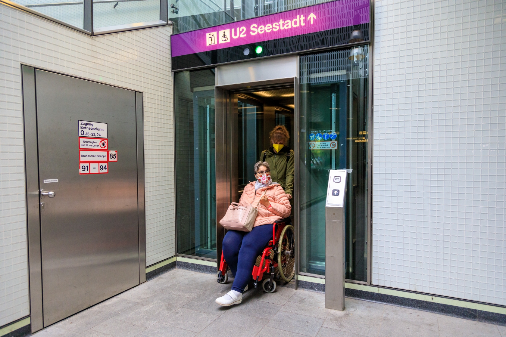 Eine Dame schiebt einen weiblichen Fahrgast im Rollstuhl aus einem Aufzug in einer U-Bahn-Station.