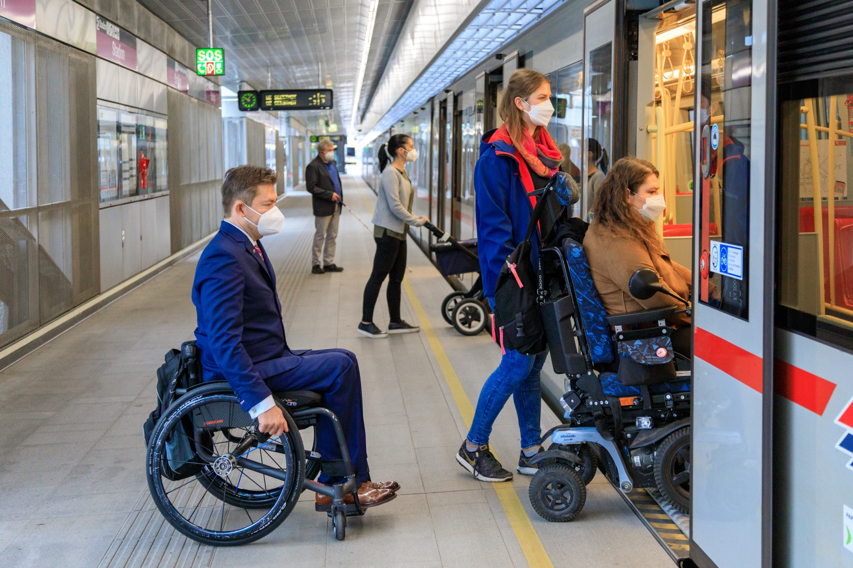 Im Vordergrund steigen drei Fahrgäste in die U-Bahn ein, zwei davon im Rollstuhl sitzend. Im Hintergrund ist eine Dame mit Kinderwagen und ein blinder Fahrgast mit Langstock beim Einstieg zu sehen.