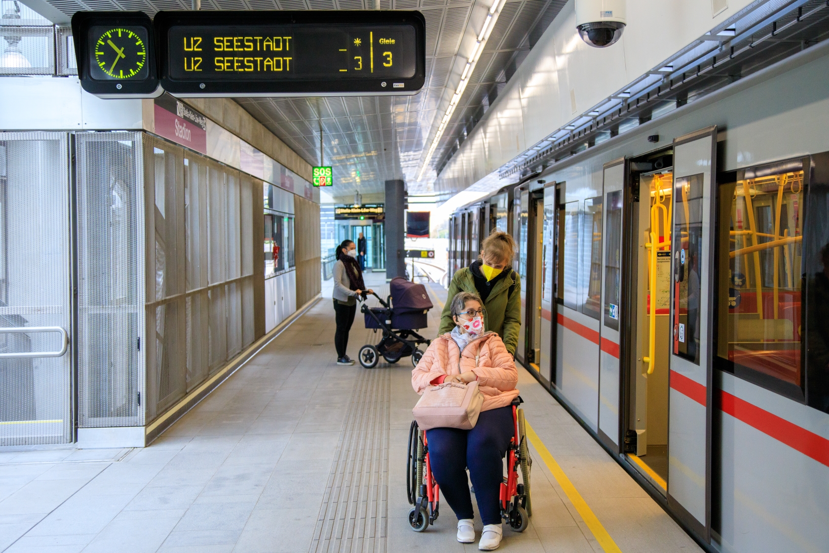 Ein-/Ausstiegsituation auf einem U-Bahnsteig. Im Vordergrund ein weiblicher Fahrgast im Rollstuhl sitzend, geschoben von einer Dame. Im Hintergrund ein weiblicher Fahrgast mit Kinderwagen. Alle tragen eine FFP2 Maske.