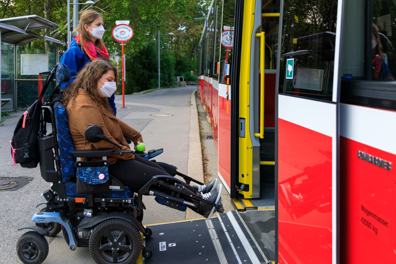 Zwei weibliche Fahrgäste, eine davon in einem elektrischen Rollstuhl, nutzen die ausklappbare Rampe, um in eine Straßenbahn einzusteigen.