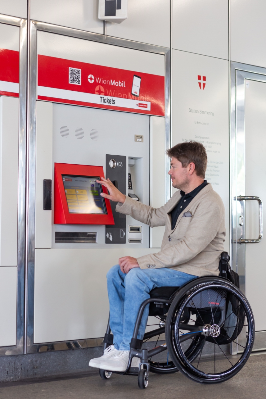 Ein Mann im Rollstuhl kauft eine Fahrkarte bei einem Fahrkarten-Automaten.